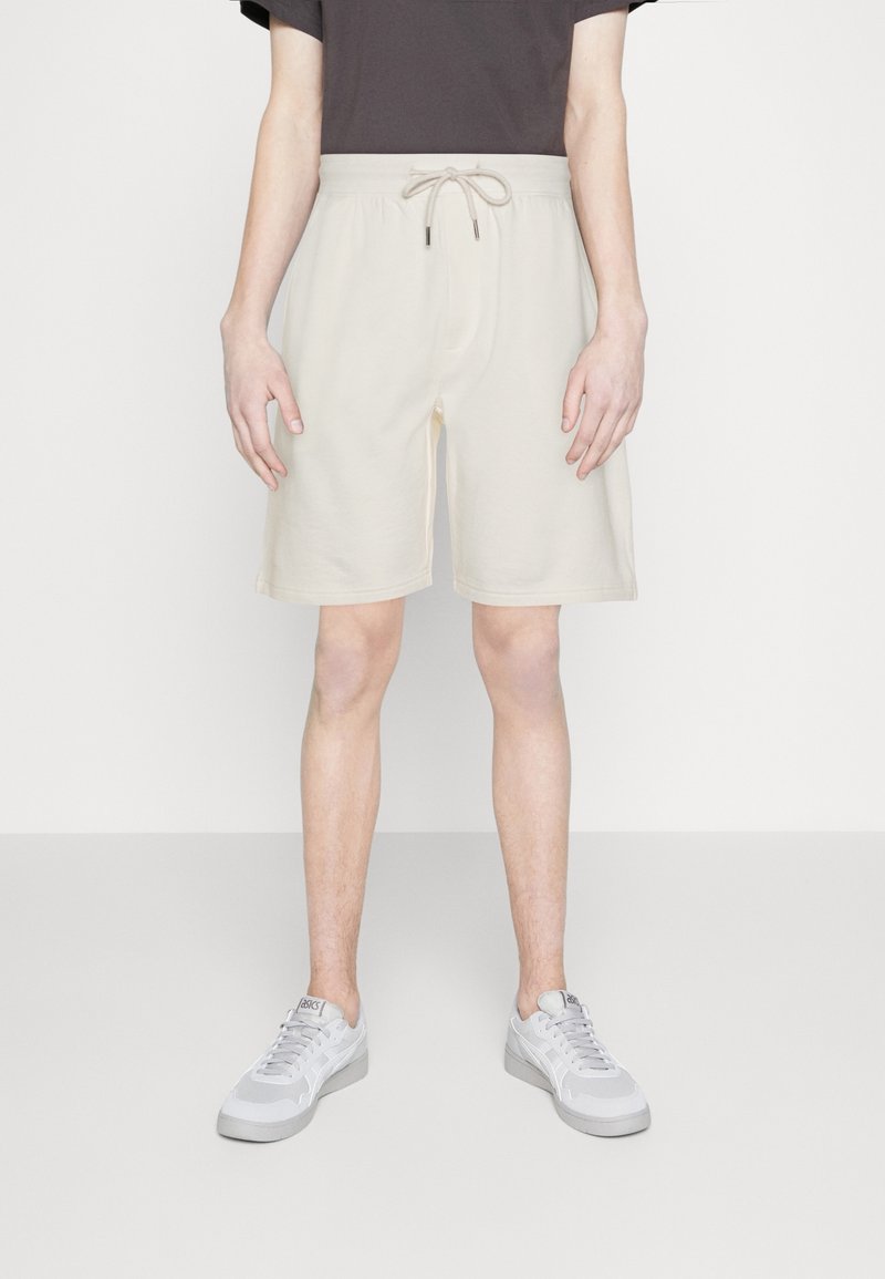 Personne portant un short beige clair à cordon, un t-shirt foncé et des baskets blanches, debout sur un sol et un fond blancs unis.