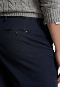 Tmavě modré kalhoty s úzkým střihem, které mají zadní kapsu s knoflíkovým zapínáním a malou logo nášivku. Tkanina má hladkou texturu.