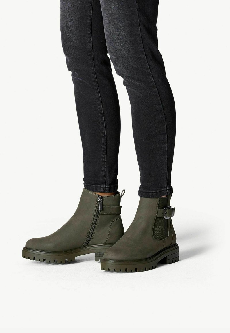 Olivově zelené kotníkové boty s hladkým, matným povrchem. Mají boční zip, detaily s přezkou a texturovanou gumovou podrážku s výrazným vzorkem.