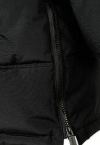 Doudoune noire avec un tissu lisse, fermeture éclair bien visible et détail de poche cousu, présentant un design épuré et minimaliste.