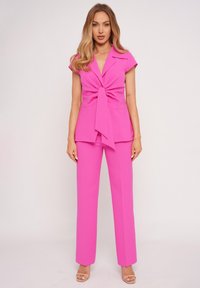 Woman standing in bright pink cap-sleeve blazer with front tie and matching trousers, wearing beige open-toe heels on plain background.