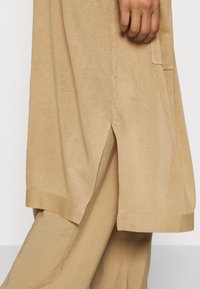 Beżowa dzianinowa tunika z bocznymi rozcięciami i nieco dłuższym tyłem, zestawiona z pasującymi beżowymi spodniami.