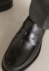 Melna ādas loafers ar gludu apdari, kuram ir dekoratīvs siksniņš pāri augšdaļai un smalkas šuves gar zoli.