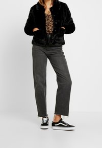 Svart fuskpälsjacka med rund krage och framfickor, bärs över en leopardmönstrad topp, tillsammans med högmidjade grå jeans och svarta sneakers.