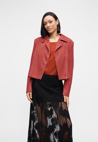 Κόκκινο μπουφάν με σχέδιο, κοντό μήκος και γιακά με σκίσιμο, συνδυασμένο με πορτοκαλί τοπ και μαύρη φούστα από δαντέλα με λουλουδένια διακόσμηση.