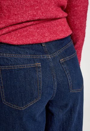 Dunkelblaue Jeans aus Denim mit orangen Nähten, ausgestattet mit zwei Gesäßtaschen und einer hohen Taille, kombiniert mit einem roten strukturierten Pullover.