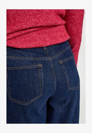 Jeans in denim blu scuro con cuciture arancioni, dotati di due tasche posteriori e una vita alta, abbinati a un maglione rosso con texture.