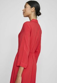 Raudona suknelė su ilgomis rankovėmis, apykakle, susiraukšlėtu liemeniu ir plazdančiu audiniu. Sklandi tekstūra ir kulkšnies ilgio dizainas. Rodoma šoninė perspektyva.