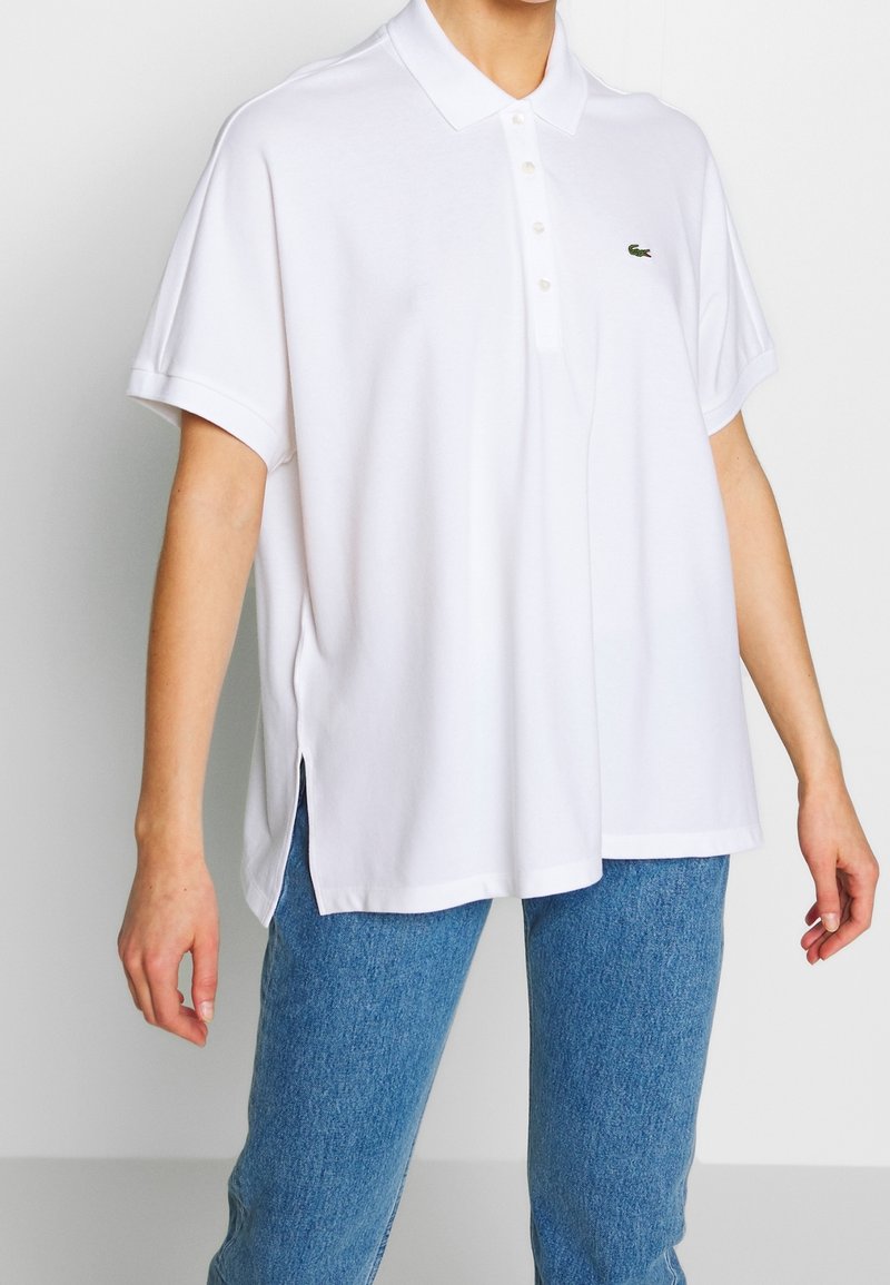 Personne portant un polo blanc ample à manches courtes et un jean bleu, le polo a un petit logo crocodile vert sur la poitrine.