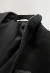 Interiorul sacoului negru are un material neted, o guler texturat și un model cu puncte subtile. Douății sunt elegante, cu un eticheta albă în contrast.