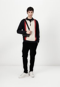 Schwarz-creamfarbener Reißverschluss-Sweatshirt mit roten Streifen und strukturiertem Vorderteil. Kombiniert mit schwarzen Cordhosen und weißen Turnschuhen, dazu eine schwarze Tasche.