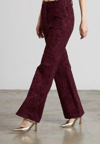 Pantalones de pierna ancha burdeos con un patrón floral texturizado, que cuentan con bolsillos laterales y una cintura alta, combinados con tacones metálicos de punta afilada.