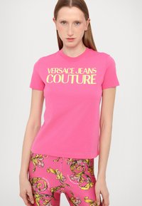 T-shirt in cotone rosa con logo giallo "VERSACE JEANS COUTURE" sul petto. Maniche corte e design classico con girocollo.
