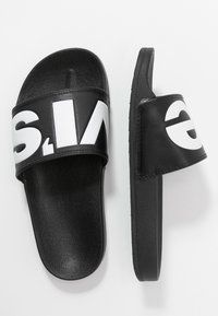 Des sandales noires à glissière en matériau synthétique texturé, avec une large sangle ornée d'inscriptions blanches, une semelle plate et un design minimaliste.