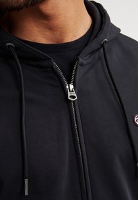Schwarze Zip-Hoodie mit glatter Textur, silbernem Reißverschluss und einem kleinen Markenpatch auf der linken Seite, mit Zugbändern zur Anpassung.