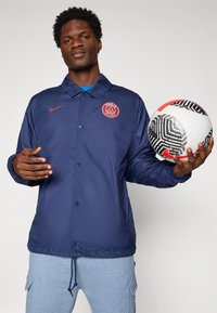 Man in navy PSG jacket and gray pants holding a white and black patterned soccer ball with red accents against a plain background.
