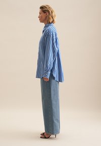 Blue and white striped oversized button-up shirt, long sleeves, paired with high-waisted blue denim wide-leg pants and black heeled sandals.