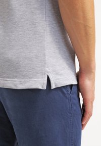 Grå texturerad polo t-shirt med sidofickor vid fållen, parat med mörkblå byxor, visar detaljer av tyg och passform.