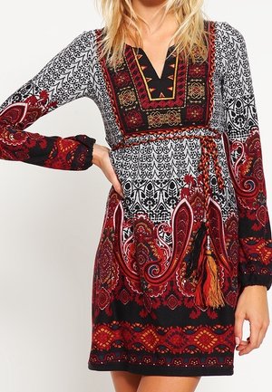 Vrouw draagt een langemouwen jurk met een patroon in rood, zwart, wit en oranje met paisley en geometrische ontwerpen, met een gevlochten riem met franjes.