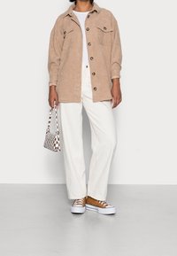 Beige Cordjacke aus Cord mit zwei Brusttaschen, weißem T-Shirt, langen weißen Hosen und braunen Sneakern mit weißen Sohlen. Taschen-Handtasche mit Schachbrettmuster.