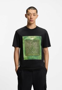 Zwarte katoenen t-shirt met een groene labyrint grafische print en de tekst "HUGO" boven het ontwerp. Korte mouwen en een losse pasvorm.