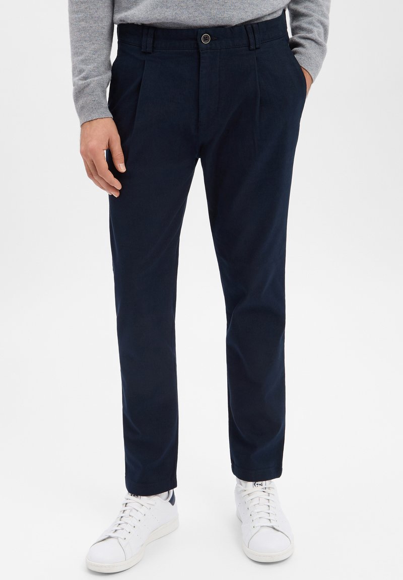 Pantalon plissé bleu marine en tissu lisse, avec une taille ajustée et un léger resserrement aux chevilles, porté avec des baskets blanches.