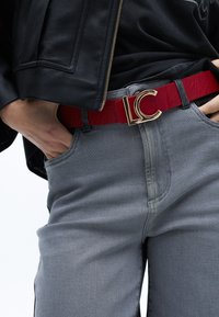 Červený lesklý pásek se zlatou sponou "LC", nošený přes šedé džíny. Kožená bunda má zip a hladkou texturu.