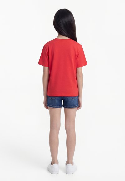Παιδί που στέκεται με την πλάτη, φοράει κόκκινο κοντομάνικο μπλουζάκι, μπλε τζιν σορτς και λευκά αθλητικά σε λευκό φόντο.