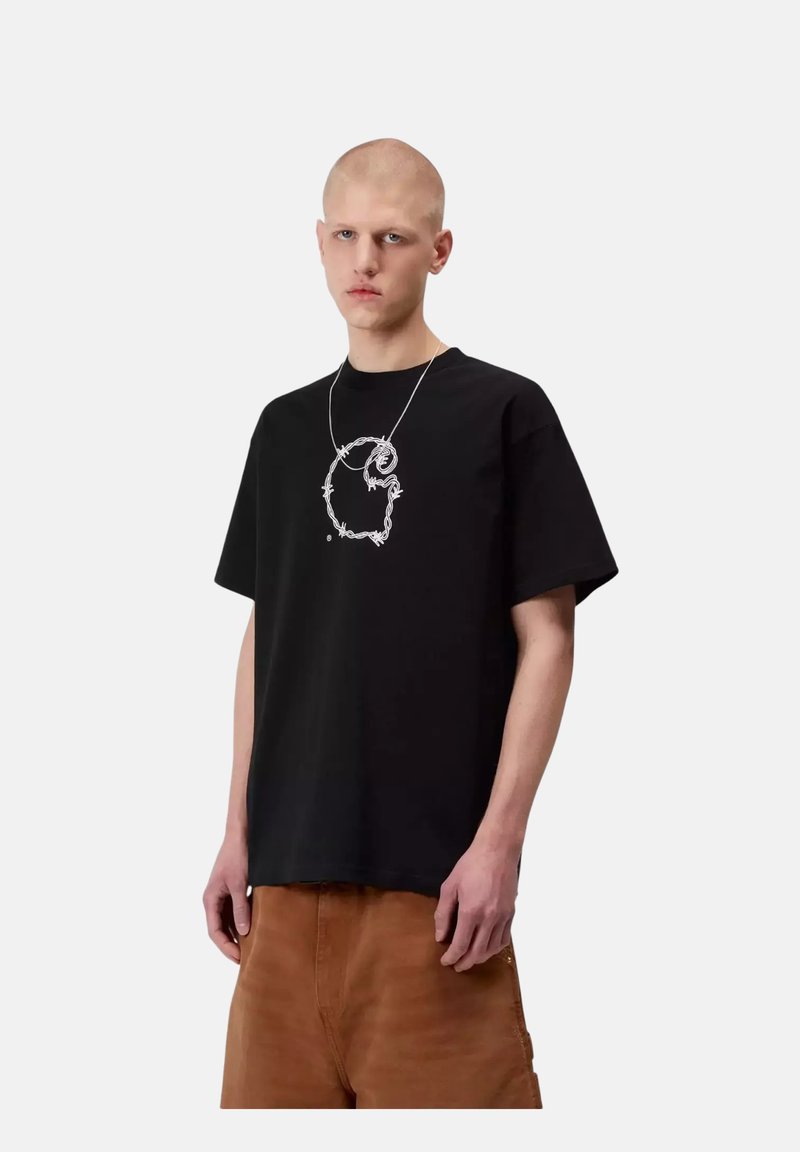 Junge Person mit rasiertem Kopf trägt ein schwarzes T-Shirt mit weißem Stacheldraht-Design und braune Hosen, steht vor schlichtem Hintergrund.