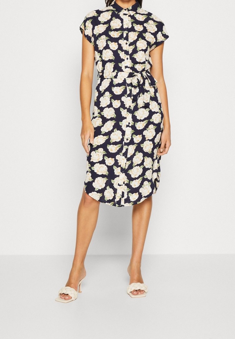 Marinblå, blommig skjortklänning med korta ärmar, knäppning fram, bälte i midjan och knälång fåll. Bärs med ljusa klacksandaler.