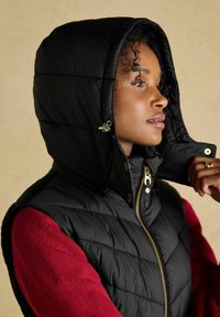 Czarna pikowana kamizelka puchowa z kapturem, podszewka z gładkiego materiału. Posiada złoty zamek i guziki, noszona na czerwonym swetrze o fakturze.