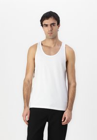 Débardeur en coton blanc avec un col rond large et une coupe décontractée, associé à un pantalon noir, présenté sur un fond uni.