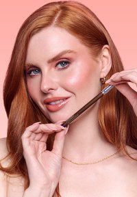 Matita eyeliner marrone dal design elegante tenuta da una persona con capelli rame di lunghezza media, pelle chiara e una sottile collana dorata.