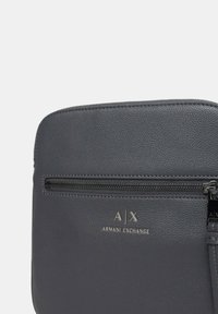 Borsa in pelle grigia scura con superficie texture, dotata di una chiusura superiore a zip e logo in rilievo "A|X ARMANI EXCHANGE". Forma rettangolare con accenti minimali.