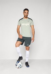 Light green athletic shirt with black accents, featuring a logo and "Bingx" text, paired with dark green shorts and white socks. Black cleats and soccer ball.