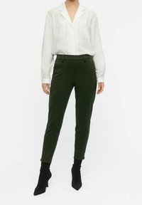Chemise blanche à manches longues avec deux poches poitrine, associée à un pantalon tapered vert foncé. Des bottines noires à bouts pointus complètent la tenue.