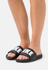 Svarta slidesandaler med breda remmar som visar ordet "LIKE" i fet vit text. Mjuk textur och platt sula.