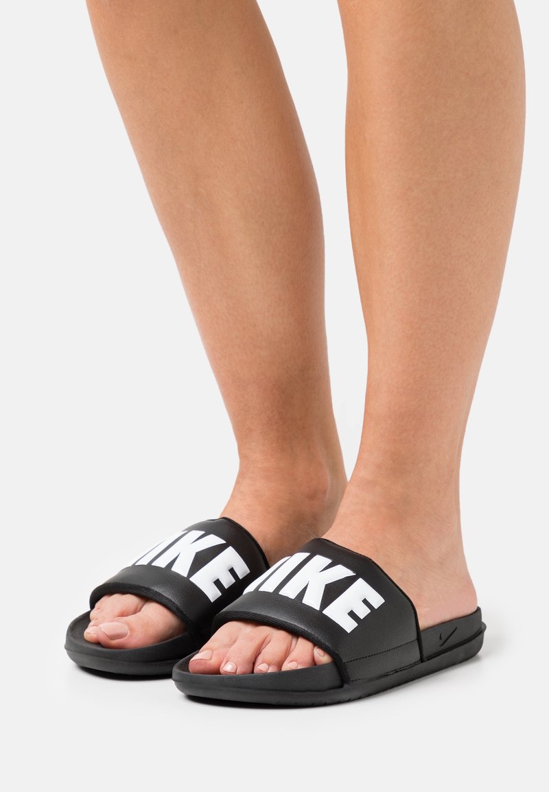 Svarta slidesandaler med breda remmar som visar ordet "LIKE" i fet vit text. Mjuk textur och platt sula.
