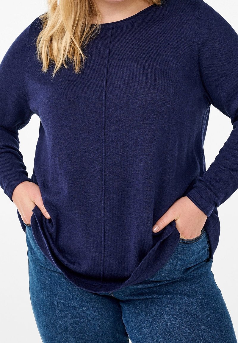 Personne portant un pull bleu foncé à manches longues et un jean bleu, les mains dans les poches, sur un fond blanc uni.
