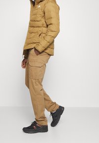 Puffjacke in Khaki, mit hohem Kragen und Reißverschlusstaschen. Kombiniert mit beige Cargo-Hosen mit einer Seitentasche. Schwarze Schuhe mit orangefarbenen Akzenten.