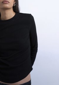 Γυναίκα που φοράει μαύρο ριμπ τοπ με μακριά μανίκια και ασημένια σκουλαρίκια κρίκους, εμφανίζεται από τους ώμους μέχρι τη μέση σε απλό ανοιχτόχρωμο φόντο.