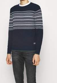 Marinblå tröja med vita geometriska mönster, rund halsringning, ribbade ärmslut och fåll med gröna inslag. Bärs med mörkgrå jeans.