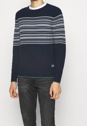 Pull marin avec des motifs géométriques blancs, col rond, manches et ourlet côtelés dotés d'accents verts. Porté avec un jean gris foncé.
