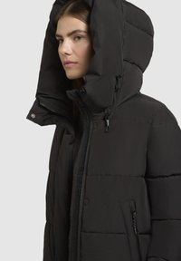 Jauna moteris vilki juodą pūkinę striukę su didele kapuce, matoma užtrauktuku ir užtrauktuku uždara šonine kišene, atsigręžusi į kairę neutraliame fone.