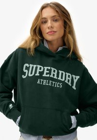 Joven mujer con una sudadera verde oscuro que dice "SUPERDRY ATHLETICS", superpuesta a una camisa de rayas azul y blanca, con las manos en los bolsillos.