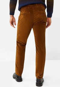 Braune Cordhosen mit vertikalen Rippungen, ausgestattet mit zwei Gesäßtaschen mit Knopfverschluss. Getragen mit schwarzen Schuhen. Nahtloses Design.