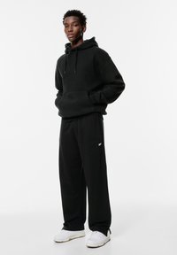 Schwarzer, übergroßer Hoodie mit Kängurutasche und Kapuze mit Zugband, kombiniert mit schwarzen, weit geschnittenen Sweatpants mit kleinem weißem Logo und weißen Sneakers.