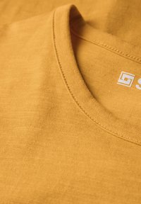 Žluté bavlněné tričko s kulatým výstřihem, s texturovanou tkaninou a bílým logem poblíž límce. Hladký povrch a rovné švy.