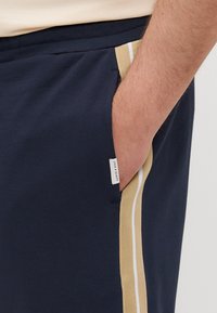 Marinblåa träningsshorts i bomullsblandning, med sidofickor och en kontrasterande beige rand samt en etikett. Mjuk textur och avslappnad passform.