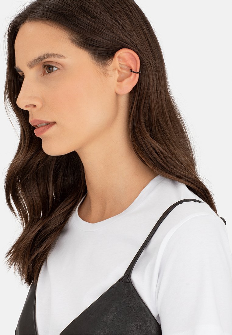 Schwarzes Ketten-Ohrcuff an einem Ohr, getragen mit einem weißen T-Shirt und einem schwarzen Trägershirt. Lange, wellige braune Haare umrahmen teilweise das Gesicht.